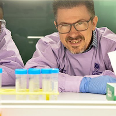 two men in purple lab coats and wearing glasses look at a rack of blue-lidded sample pots